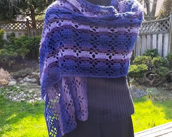 May include: A blue and purple crochet shawl, displayed on a mannequin. The shawl has an openwork design, with alternating solid and patterned sections. The garment is shown outdoors, with a blurred background of trees and a wooden fence.