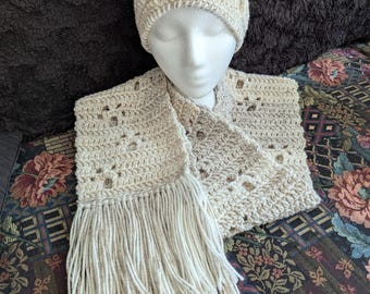 May include: A cream-coloured crochet hat and scarf set. The hat is a beanie style with openwork details. The scarf is rectangular with fringe at the ends. The set is displayed on a mannequin head, showcasing the handmade texture and design.