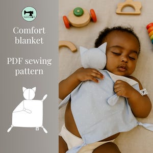 Op de afbeelding: Een slapende baby met een blauw-witte knuffeldekentje en een katvormig knuffel. Het dekentje heeft een stippenpatroon en gestreepte randen. Houten speelgoed ligt rond de baby. De tekst "Comfort blanket PDF sewing pattern" is ook zichtbaar.