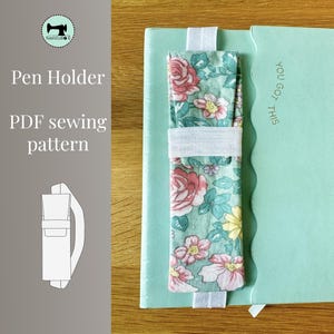 May include: A mint green notebook with the text "You Got This" and a floral pen holder. The pen holder is made of fabric with a pattern of pink roses, yellow flowers, and green leaves. A white elastic band secures the pen holder to the notebook.