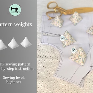 May include: Three white pyramid-shaped sewing weights and four patterned fabric sewing weights on a white fabric with a polka dot pattern. The fabric is on top of a white sewing pattern. The image also includes a pair of gold scissors.