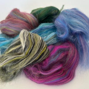 May include: A selection of dyed fibre for crafts, in shades of pink, blue, green, and yellow. The fibres are soft and fluffy, possibly wool or a similar material. The fibres are arranged in a circular shape, ideal for spinning or felting projects.