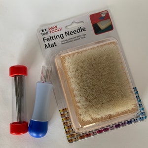 May include: A set of felting tools including a felting mat, a needle tool, and a case of felting needles. The felting mat is made of a light brown material with a short, dense pile. The needle tool is blue and white with a red tip. The needle case is clear plastic with a red top and bottom. The packaging has the text "WoW TOOLS Felting Needle Mat" and "SUITABLE FOR USE WITH FTO1 Wow FELTING NEEDLE TOOL".