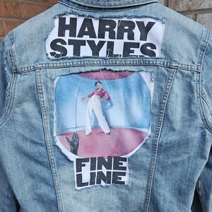 May include: Light blue denim jacket with patches. The top patch reads "HARRY STYLES" in bold white letters. Below is a colorful graphic of a person in a pink shirt and white pants. The bottom patch reads "FINE LINE".