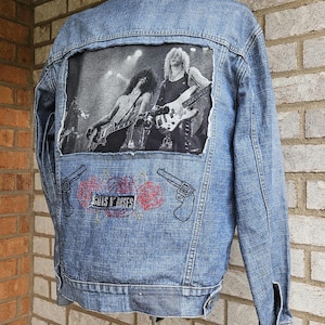 May include: A light blue denim jacket featuring a black and white band image on the back. Embroidered below the image are the words "GUNS N' ROSES", two guns and roses.