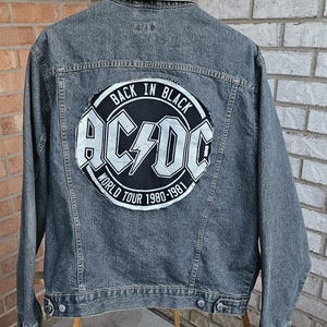 May include: A gray denim jacket with a large, circular patch on the back featuring the AC/DC logo and the text "BACK IN BLACK WORLD TOUR 1980-1981". The jacket is button-down with two chest pockets and is displayed on a wooden hanger.