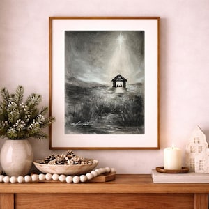 May include: A framed black and white print of a nativity scene. The artwork depicts a small stable illuminated by a bright star, set in a field. The print is displayed on a wooden table with decorative items.