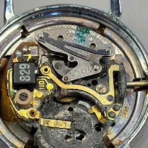 May include: Close-up of a watch mechanism with a silver-colored case. The internal components include gold-colored gears, a black circuit board with the number "829", and the brand name "TIMEX". The watch is disassembled, revealing its intricate workings.