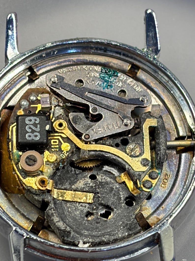 May include: Close-up of a watch mechanism with a silver-colored case. The internal components include gold-colored gears, a black circuit board with the number "829", and the brand name "TIMEX". The watch is disassembled, revealing its intricate workings.