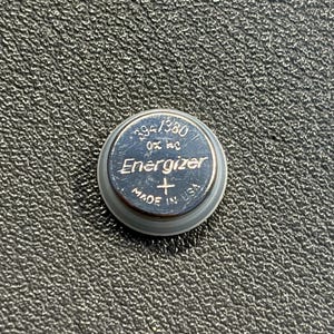 May include: A silver Energizer 394/380 button cell battery. The battery has the text "Energizer" and "Made in USA" printed on it. The battery is on a textured black surface.