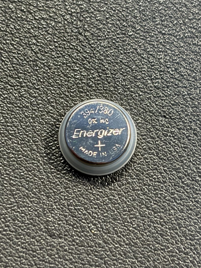 May include: A silver Energizer 394/380 button cell battery. The battery has the text "Energizer" and "Made in USA" printed on it. The battery is on a textured black surface.