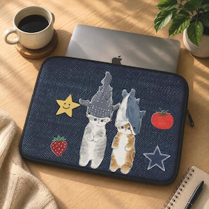 May include: A dark blue denim-textured laptop sleeve featuring two cats wearing shark hats, a yellow smiling star, a red strawberry, a red tomato, and a dark blue star outline. The sleeve is rectangular with a black zipper along the edge.