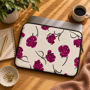 Può includere: Una custodia rettangolare per computer portatile con sfondo color crema e un motivo ripetuto di fiori astratti color magenta, con sottili steli e contorni neri disegnati a mano. La custodia presenta una chiusura a zip nera sul bordo.