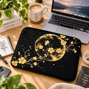 May include: A black laptop sleeve featuring a large, golden full moon graphic overlaid with delicate, blooming cherry blossom branches in a metallic gold tone. The design includes scattered gold specks across the dark background.