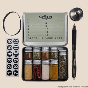 May include: A metal spice tin with a green label that says "SPICE UP YOUR LIFE". The tin is open and contains 8 small glass jars with various spices. The jars are labelled with numbers 1 through 8. The tin also has a set of numbered stickers on the side.