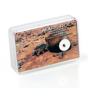 May include: A clear plastic display case containing a piece of Mars rock. The case features an image of the Martian surface with a rover and a large rock. Text on the case reads "MARS ROCK" and details about the meteorite.
