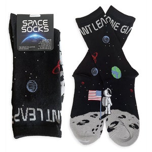May include: A pair of black socks with a space theme. One sock features an astronaut standing on the moon with the American flag, the other sock has a planet and stars. The socks have the text "Giant Leap One Giant" on the cuff.