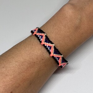 May include: A black friendship bracelet with a geometric pattern in pink, white, and blue.