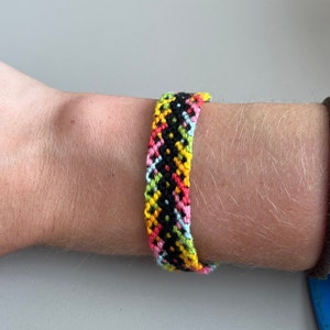May include: A colorful friendship bracelet with black, yellow, pink, green, and light blue threads woven in a geometric pattern.