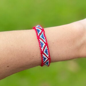 May include: A woven friendship bracelet with a red border and a repeating pattern of blue and white diamonds. The bracelet is worn on a person's arm against a blurred green background.