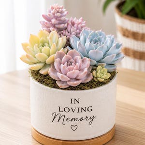 May include: A white cylindrical ceramic planter with a wooden base containing a variety of colorful succulents in shades of pastel pink, yellow, and blue. The planter features the black text IN LOVING Memory and a small heart symbol on the front.