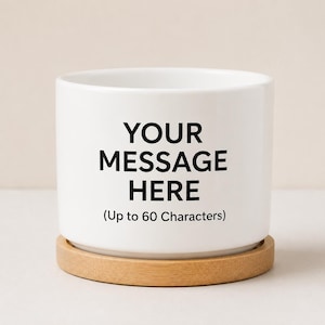 May include: A white ceramic cylindrical planter with a smooth finish, resting on a circular light-colored wooden base. The front features black text that reads: YOUR MESSAGE HERE (Up to 60 Characters). The design is simple, clean, and functional.