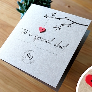 May include: A gray card with the text "to a special dad" and "Happy Birthday 80" in black script. A red heart with two white buttons is hanging from a branch with leaves.