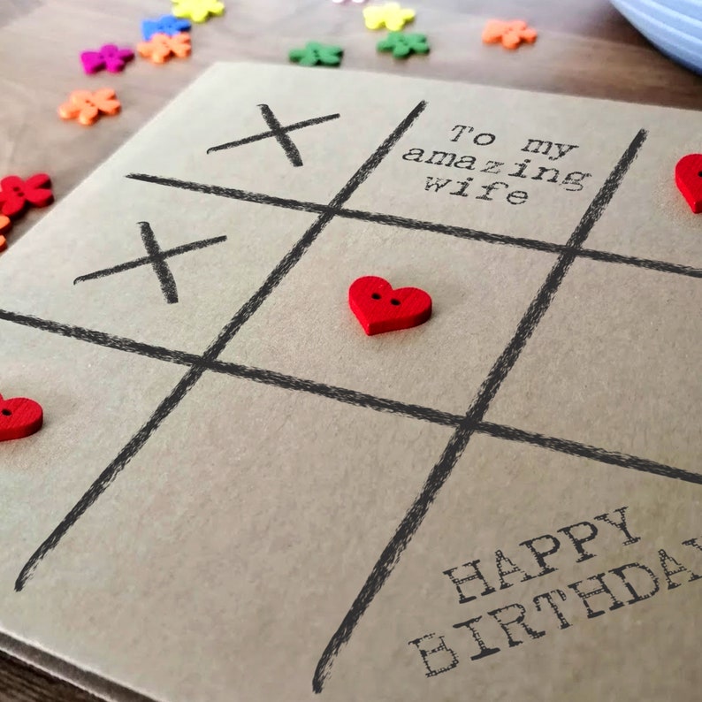 May include: A brown card with a tic-tac-toe game drawn on it. The game is played with red heart buttons. The card says "To my amazing wife" and "Happy Birthday".