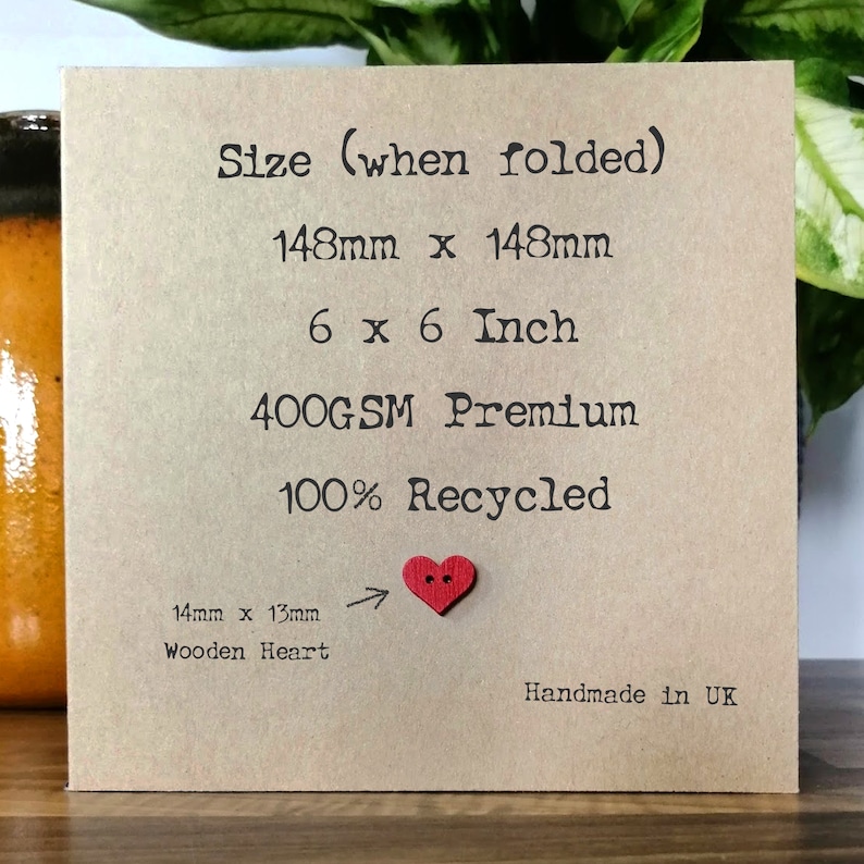 May include: A brown card with the text "Size (when folded)" and "148mm x 148mm", "6 x 6 Inch", "400GSM Premium", "100% Recycled", "14mm x 13mm Wooden Heart", and "Handmade in UK". A small red wooden heart is attached to the card.