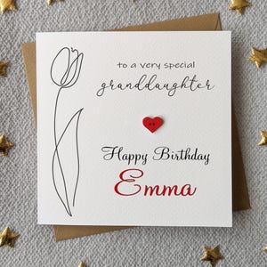 May include: A white birthday card with a brown envelope. The card features a black line drawing of a tulip, the text "to a very special granddaughter", a red heart-shaped button, and "Happy Birthday Emma" in red.