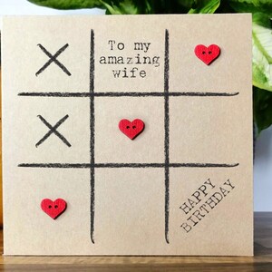 May include: A brown card with a black tic-tac-toe board and red heart buttons. The card says "To my amazing wife" and "Happy Birthday".