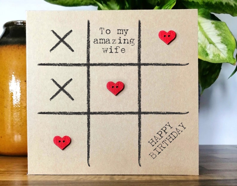 May include: A brown card with a black tic-tac-toe board and red heart buttons. The card says "To my amazing wife" and "Happy Birthday".