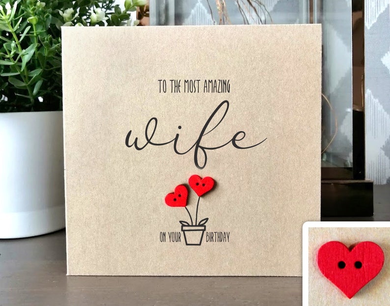 May include: A square, tan greeting card with the text "TO THE MOST AMAZING wife ON YOUR BIRTHDAY". The card features a drawing of a flower pot with two red heart-shaped flowers.