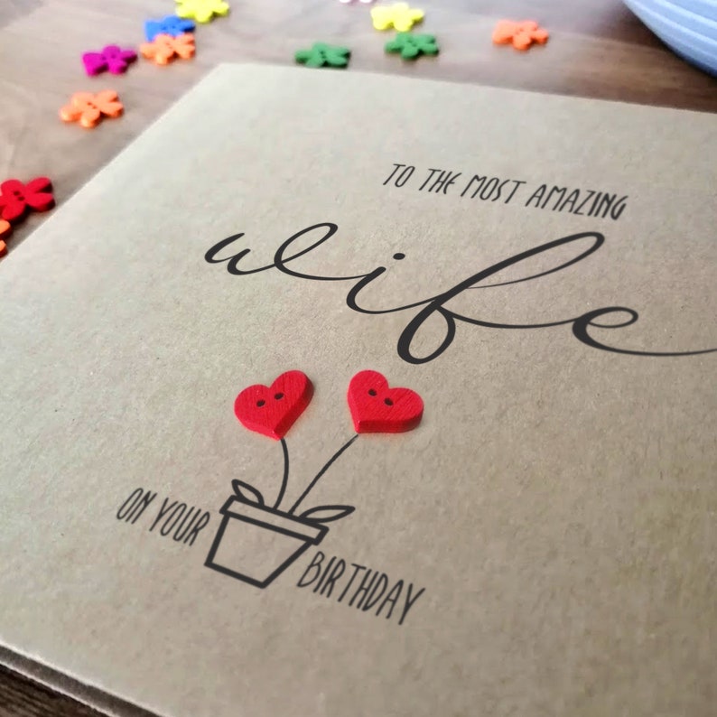 May include: A handmade birthday card with a kraft paper background. The card features the words "To the most amazing wife" in elegant script. Two red heart-shaped buttons are attached to the card, with a drawing of a flower pot below. The words "On your birthday" are printed below the flower pot.