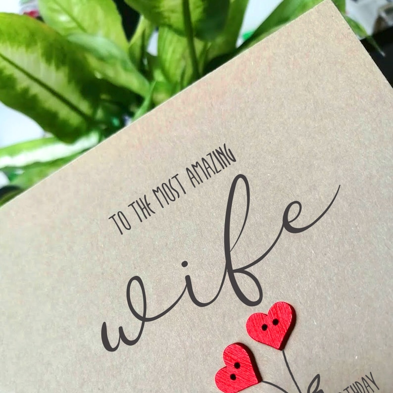 May include: A greeting card with the text "TO THE MOST AMAZING wife" in black script. Two red heart-shaped embellishments are at the bottom. The card is a light brown color.