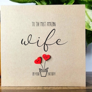 May include: A square greeting card with a kraft paper finish. The card features the text "TO THE MOST AMAZING wife ON YOUR BIRTHDAY" in black script. Two red heart-shaped embellishments are placed above a small pot.