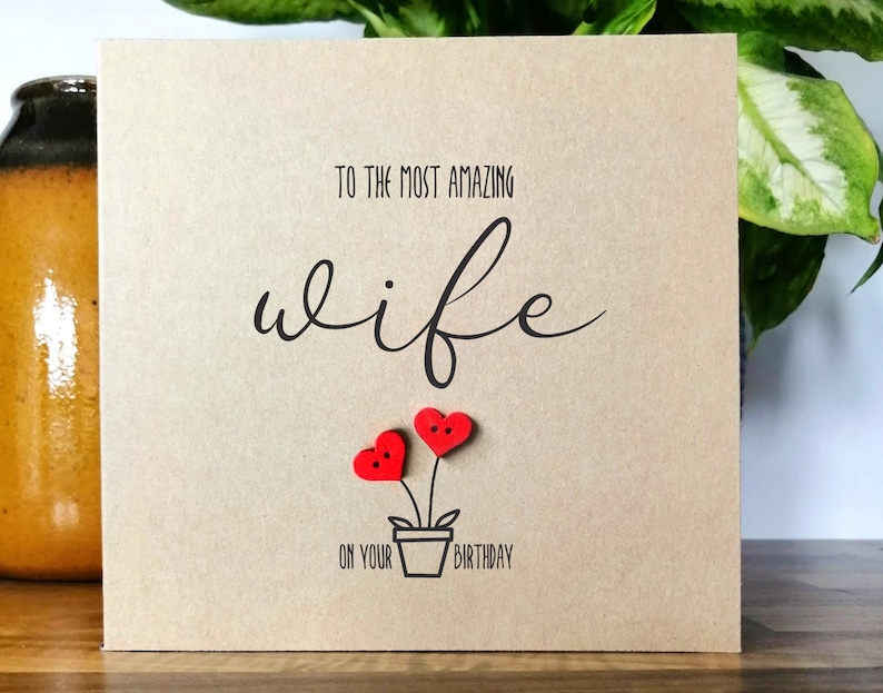May include: A square greeting card with a kraft paper finish. The card features the text "TO THE MOST AMAZING wife ON YOUR BIRTHDAY" in black script. Two red heart-shaped embellishments are placed above a small pot.