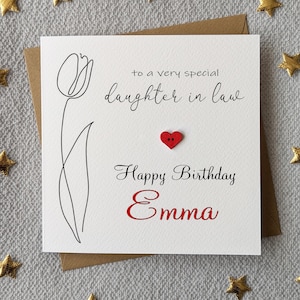 May include: A white birthday card for a daughter-in-law, with a black tulip outline and the text "to a very special daughter in law." A red heart-shaped button is above "Happy Birthday Emma" in red script. The card is on a brown envelope.