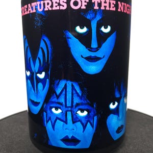 May include: Black cylindrical tumbler featuring the faces of four people with blue face paint and the text "CREATURES OF THE NIGHT" in pink. The faces have white glowing eyes. The tumbler is on a dark surface.