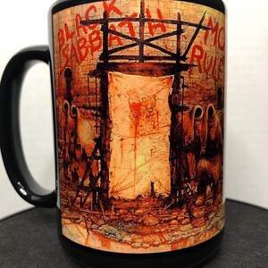 May include: Black ceramic mug featuring artwork with the words "BLACK SABBATH" and "MO RULES" in red. The artwork depicts a scene with figures and a structure against a brick wall. The mug has a black handle and rim.