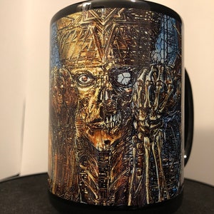 May include: Black ceramic mug with a graphic of a skeletal figure with a golden face and a blue eye. The figure is surrounded by a geometric pattern.