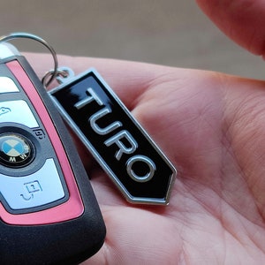May include: A black and silver key chain with the word "TURO" in white lettering. The keychain is attached to a black and silver car key with a BMW logo.