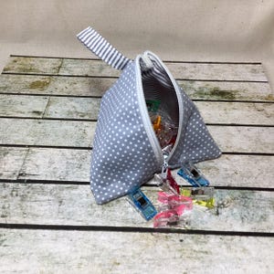 May include: A grey polka dot triangular pouch with a white zip and a striped handle. The pouch is open, revealing colourful sewing clips. The background is a wooden surface.