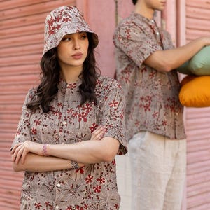 Könnte beinhalten: Zwei Personen tragen beige Hemden mit rotem Blumendruck. Die Person links trägt einen braunen und roten Bucket-Hut. Die Person rechts hält ein grünes und orangefarbenes Kissen.