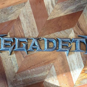 May include: A metallic gray "MEGADETH" logo, with stylized, sharp-edged lettering, rests on a wooden surface with a chevron pattern. The logo's design is three-dimensional, casting subtle shadows.