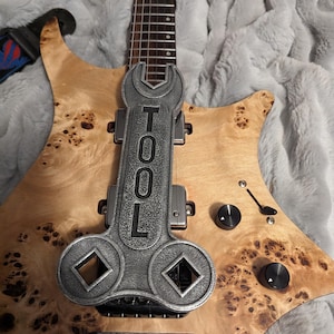 May include: A light-colored electric guitar with a unique wood grain pattern. A metal tool-shaped object is mounted on the guitar, with the word "TOOL" engraved on it. The guitar has black knobs and a strap.