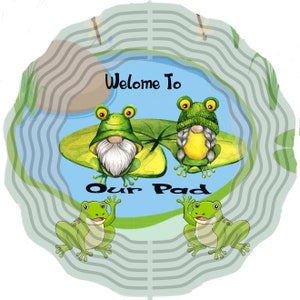 May include: Welcome to Our Pad sign with two green frogs wearing hats sitting on lily pads in a blue pond. Two more green frogs are sitting on the edge of the pond.