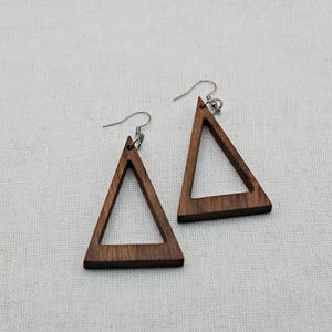 May include: A pair of geometric earrings. Each earring features a dark brown, open triangle shape made of wood, suspended from a silver-colored hook. The earrings are displayed on a light-colored, textured surface.
