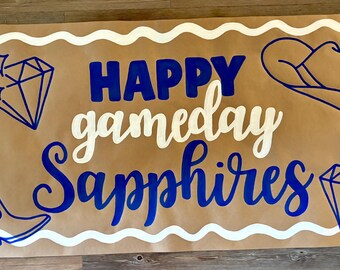 Op de afbeelding: Een bruine banner met de woorden "HAPPY gameday Sapphires" in blauw en wit. De banner heeft een witte golvende rand en bevat illustraties van een cowboyhoed, een cowboylaars en twee diamanten.