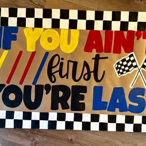 May include: A brown banner with the text "IF YOU AIN'T FIRST YOU'RE LAST" in various colors. The banner has a black and white checkered border and two checkered flags.